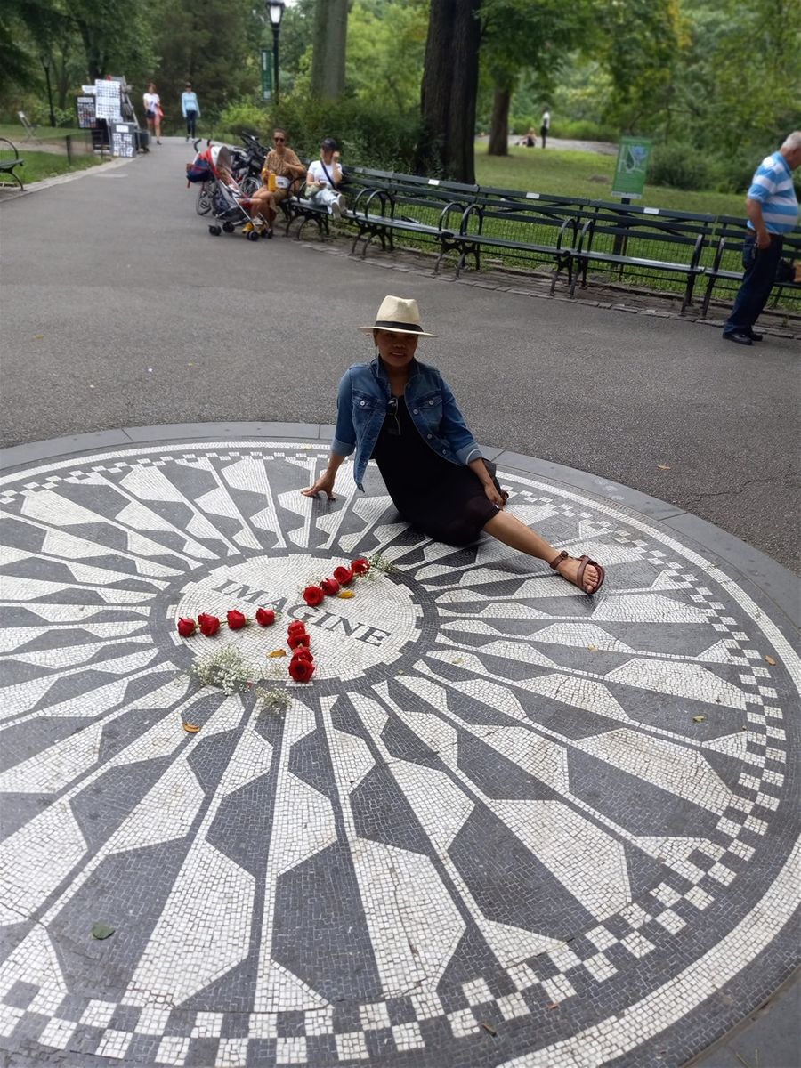 Farida at the John Lennon Memorial in Central Park NYC. I posted this photo at this time because I felt bad about the meager donations I was making to help the Ukrainians while Russia was attacking them. I thought that I could post a couple of photos from this place because it is a reminder that it is possible for all of us to live together in a better way. I posted a different photo of the John Lennon Memorial on my Arrived in New York City page. I also posted a video from here on my video page. 🙂 . This memorial is very close to the Dakota Condos Building where John Lennon and Yoko Ono lived. I believe Yoko Ono still lives there.